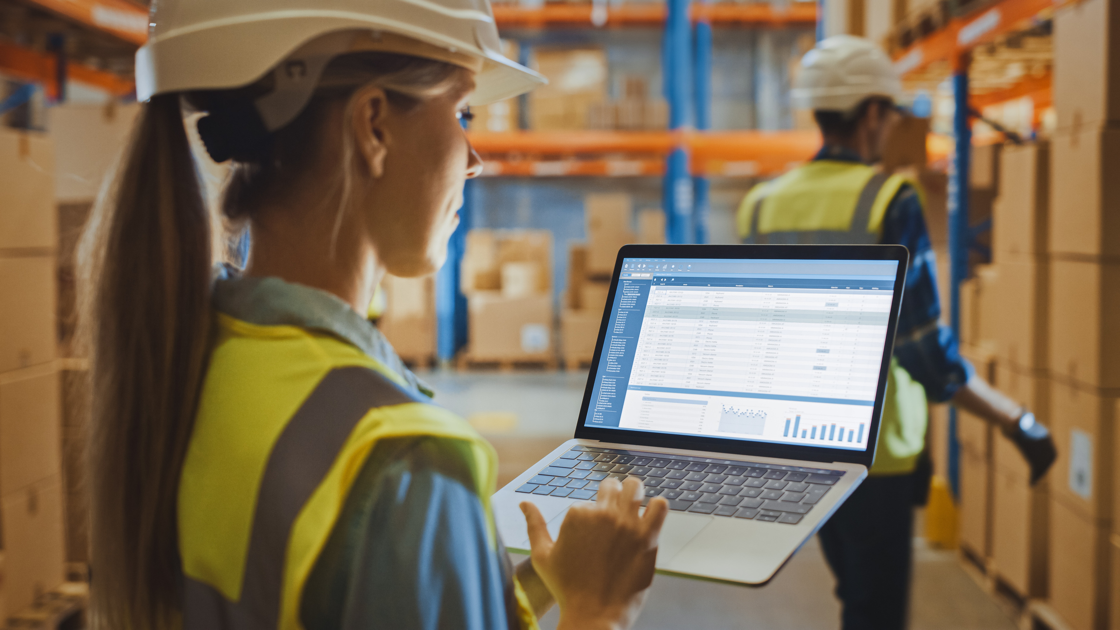 Professional Female Worker Wearing Hard Hat Holds Laptop Computer with Screen Showing Inventory Checking Software in the Retail Warehouse full of Shelves with Goods.