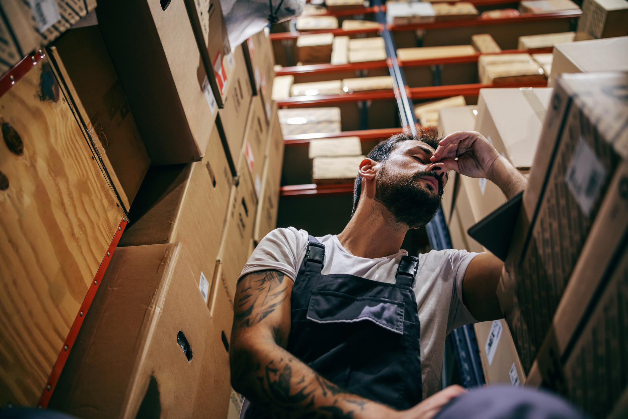 Tattooed bearded worker in overalls, overworked. Storage of import and export firm interior.
