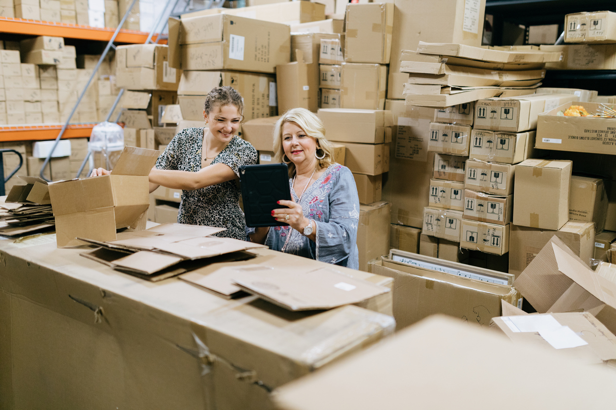 A mature woman process new product from boxes in the back of her store, taking inventory of supplies using a digital touchscreen tablet. An employee helps with the work. Preparation for stocking shelves with new supplies.