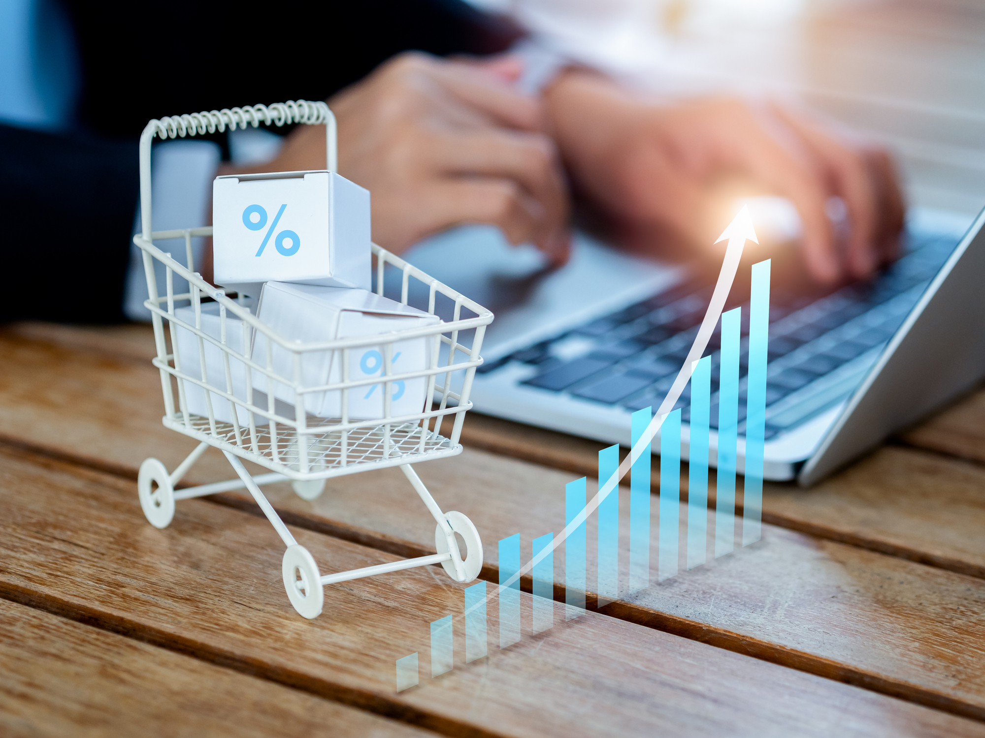 Online shopping purchase, sales increase, market trends concepts. Rising arrow on business growth graph near the shopping cart trolley with parcel boxes on desk near person using laptop computer.