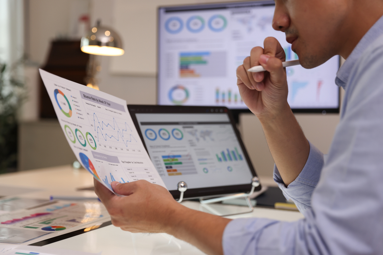 Focused businessman analysing marketing reports on a desktop monitor and reviewing paper graphs, financial stats, and startup project infographics.